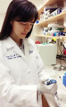 A photo of Ying Liu in a lab coat from the waist up. She is focused and looking down at her work where she is holding a pipettor inserted into a petri dish.